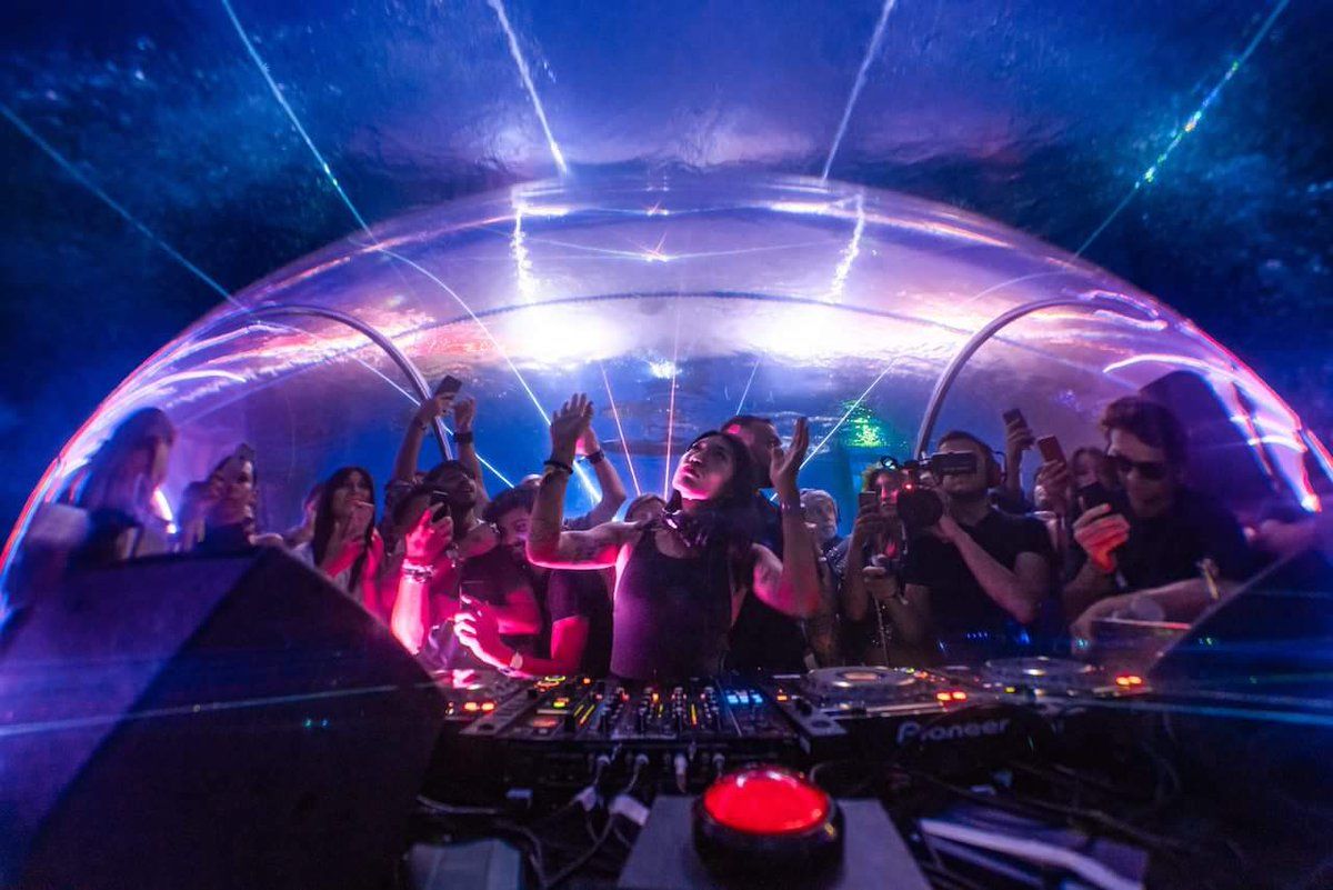 A DJ performing inside a transparent dome beneath a pool, surrounded by a crowd holding up phones, with laser beams cutting through the water above